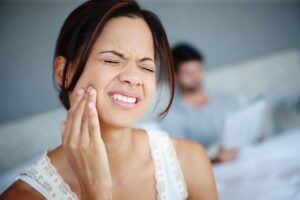 adult-woman-experiencing-tooth-pain-in-home-settin-2026-03-25-04-38-36-utc - Encino Gentle Dental Woman grimacing in pain while touching her cheek, likely a toothache, with a blurred man in the background reading a document.