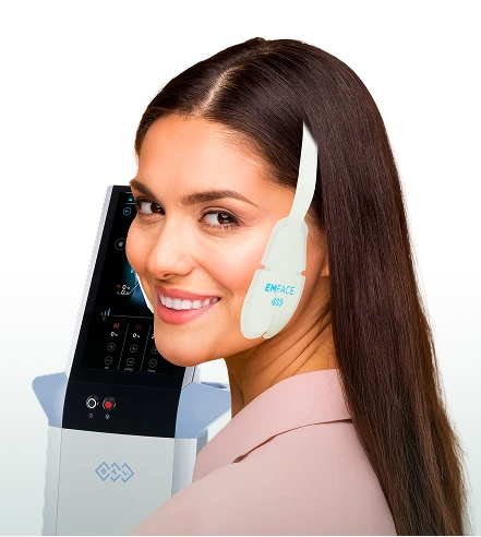 Smiling woman wearing a white facial device on her cheek, standing beside a medical machine in a clinical setting.