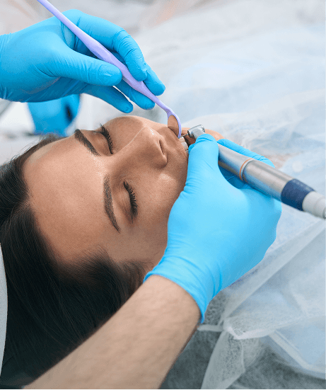 Dental procedure underway: dentist in blue gloves uses a drill and suction near a patient’s mouth while the patient rests in a dental chair.